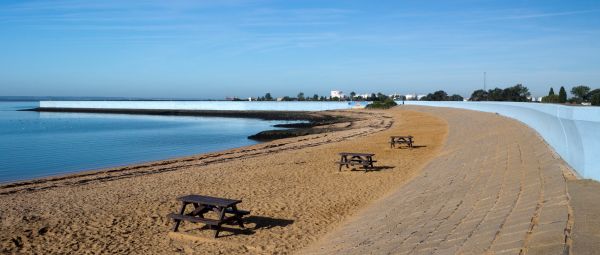 Looking out over a sandy beach, dotted with three wooden picnic tables, with a light blue sea wall bordering the sand at the back of the beach.