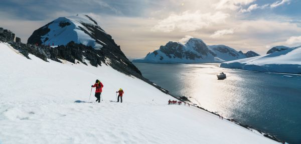 Walkers hiking up a snowy mountain in the sunshine above the sea with a ship and mountains in the background.