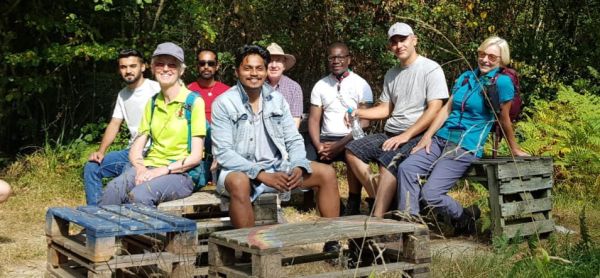 A group of asylum seekers and walk leaders smiling, standing on a bridge in a sunny woodland. 