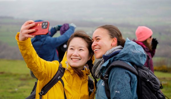 Two young walkers in rain jackets smile into a phone whilst taking a selfie.