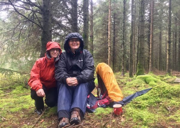 Two walkers wearing rain jackets and trousers, sitting on damp forest grass surrounded by huge trees 