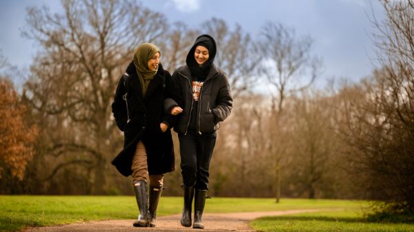 A mother and daughter walking on a flat concrete path with large trees in the background 