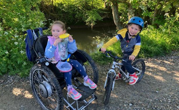 A young girl in a wheelchair on a path with her brother on a bicycle.